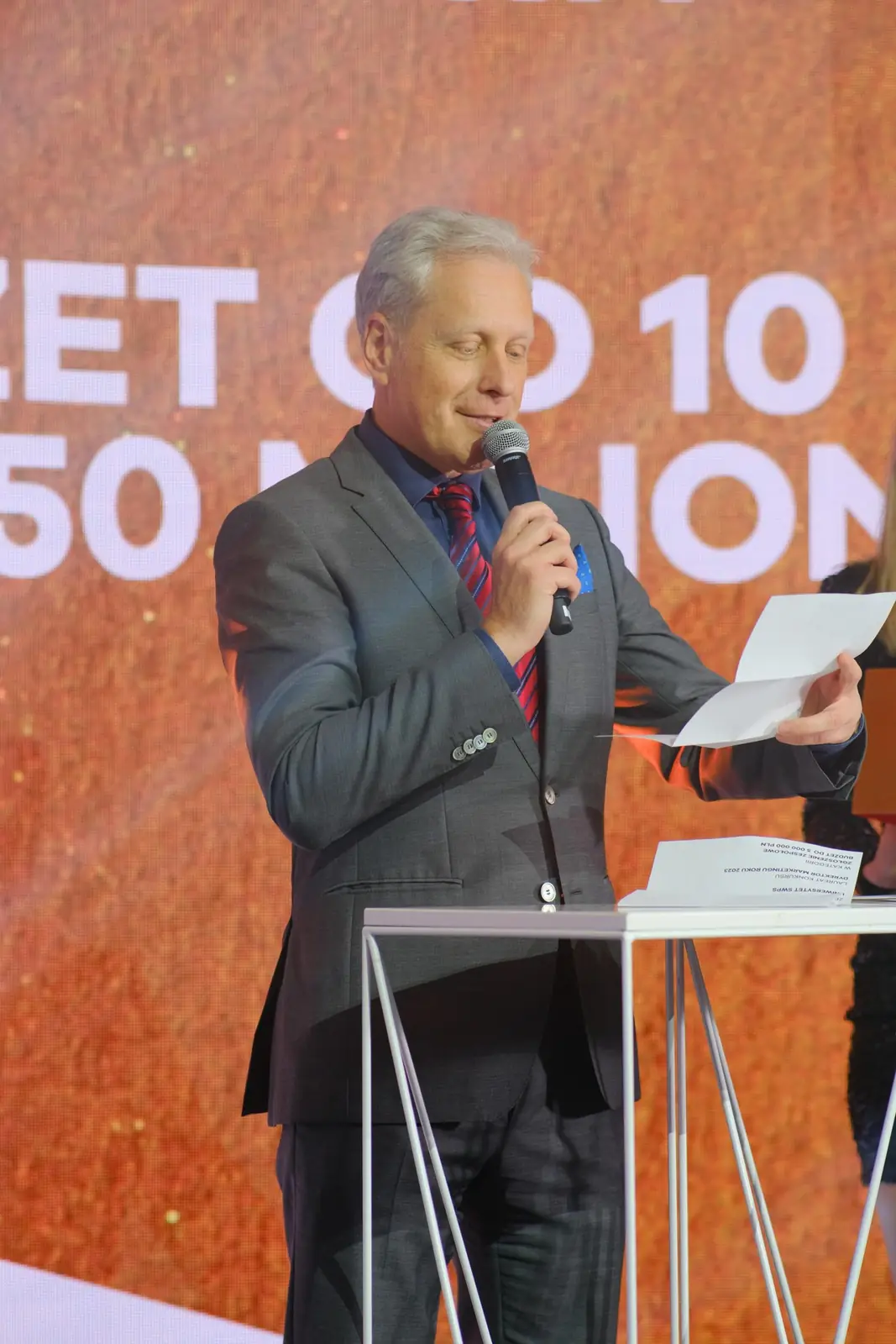 Mężczyzna w szarym garniturze odczytuje przy mównicy werdykt w kategorii budżetowej — Gala DMR XII (2023)