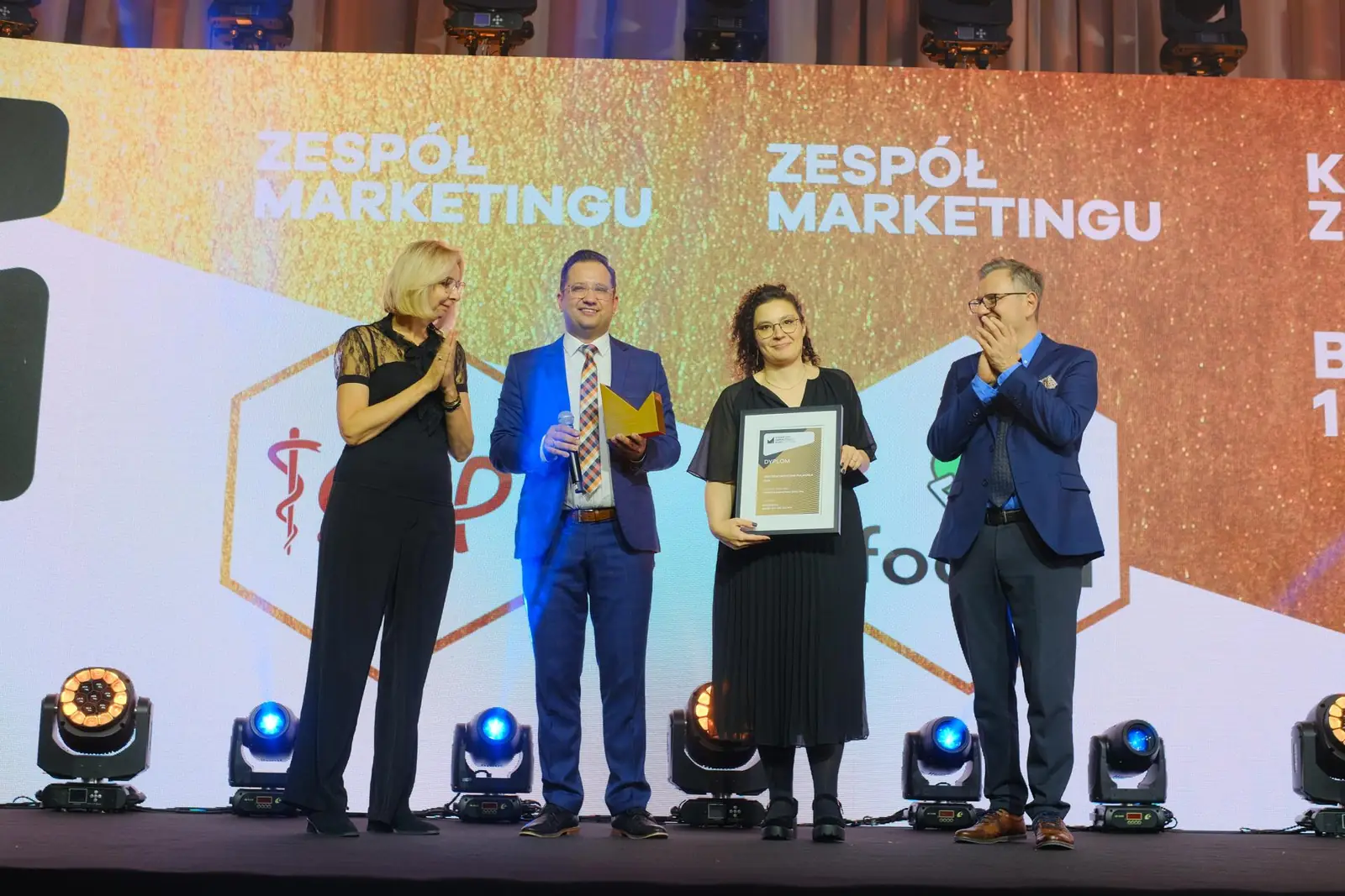 Laureaci ze statuetką i oprawionym dyplomem, a wręczający nagrodę biją im brawo na scenie — Gala DMR XII (2023)
