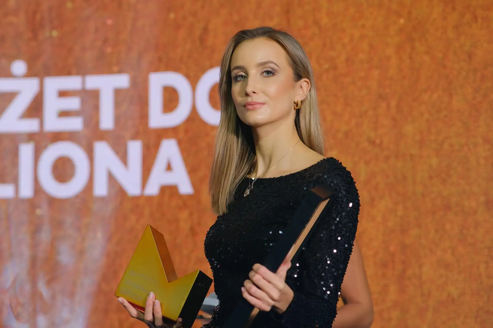 Laureatka w czarnej cekinowej sukience pozuje ze złotą statuetką na tle pomarańczowego ekranu — Gala DMR XII (2023)