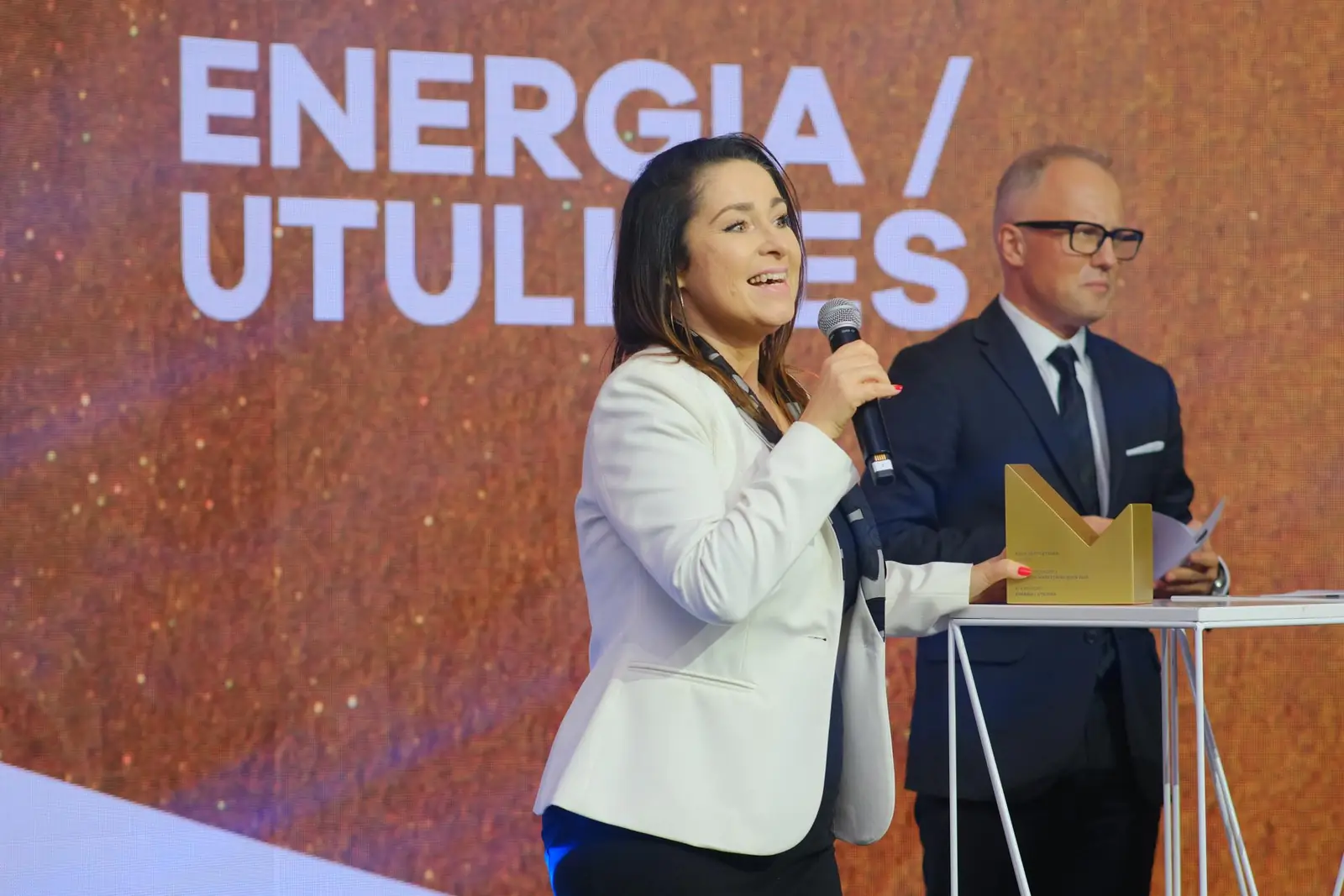 Laureatka w białej marynarce z uśmiechem przemawia, obok mężczyzna w okularach i statuetka na mównicy — Gala DMR XII (2023)