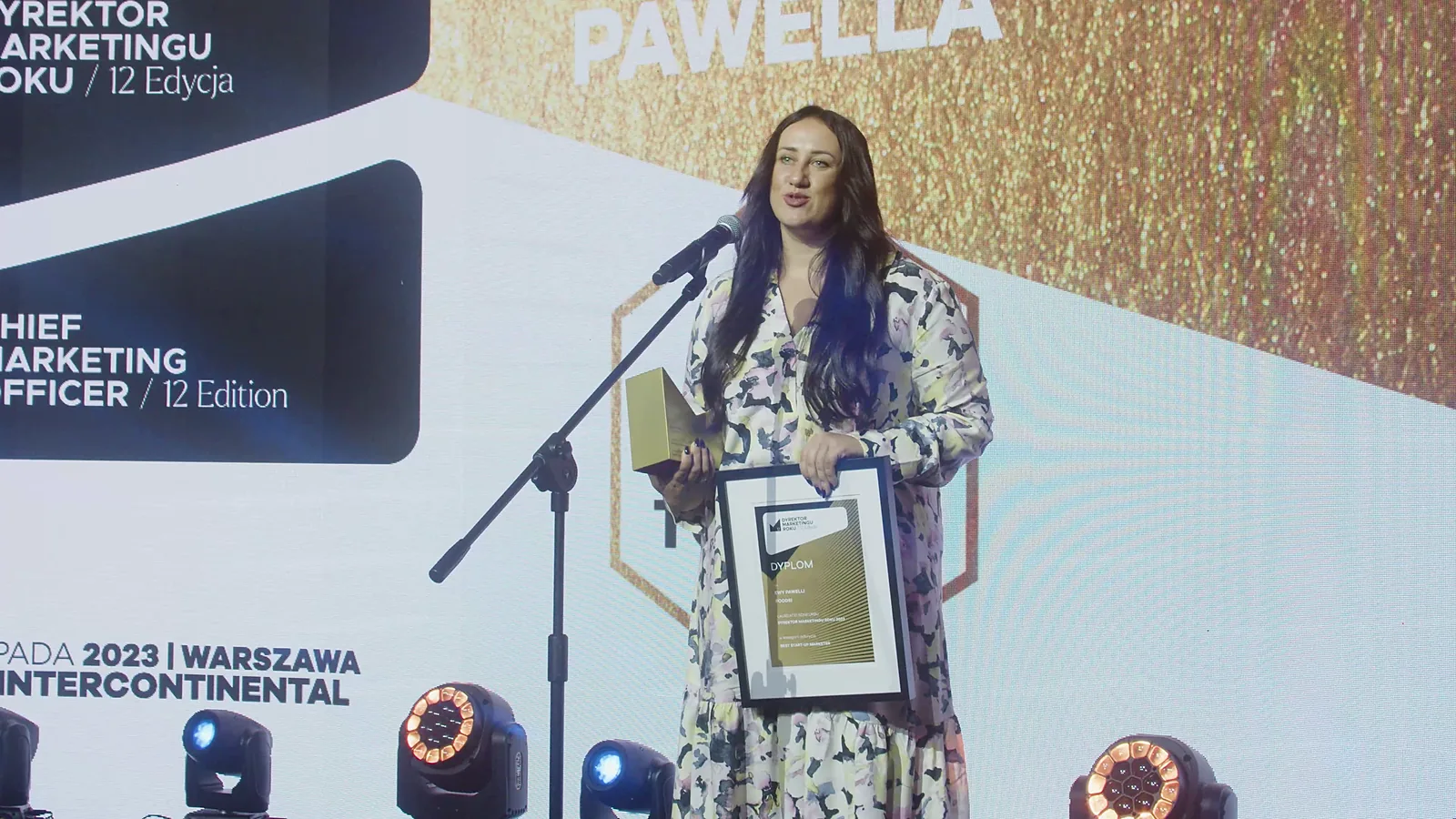 Laureatka z dyplomem i statuetką mówi do mikrofonu na statywie przy logo konkursu — Gala DMR XII (2023)