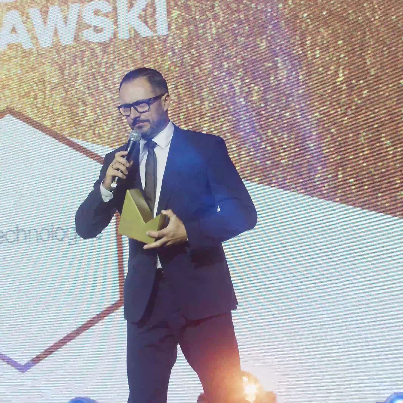Laureat w okularach, widoczny w całej sylwetce, przemawia do mikrofonu ze złotą statuetką — Gala DMR XII (2023)