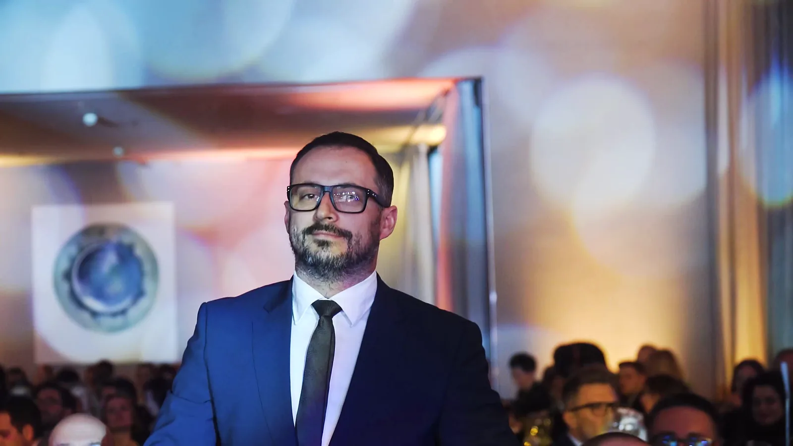 Brodaty mężczyzna w okularach i granatowym garniturze stoi wśród publiczności na tle rozmytych świateł — Gala DMR XII (2023)