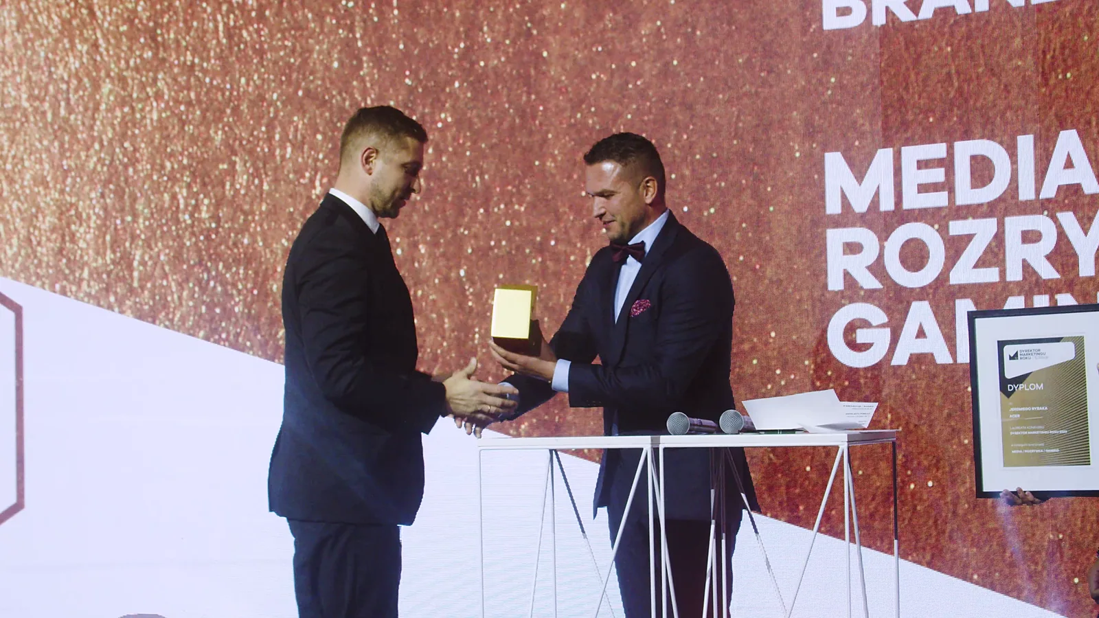 Prowadzący w smokingu wręcza statuetkę laureatowi w kategorii media, rozrywka i gaming — Gala DMR XII (2023)