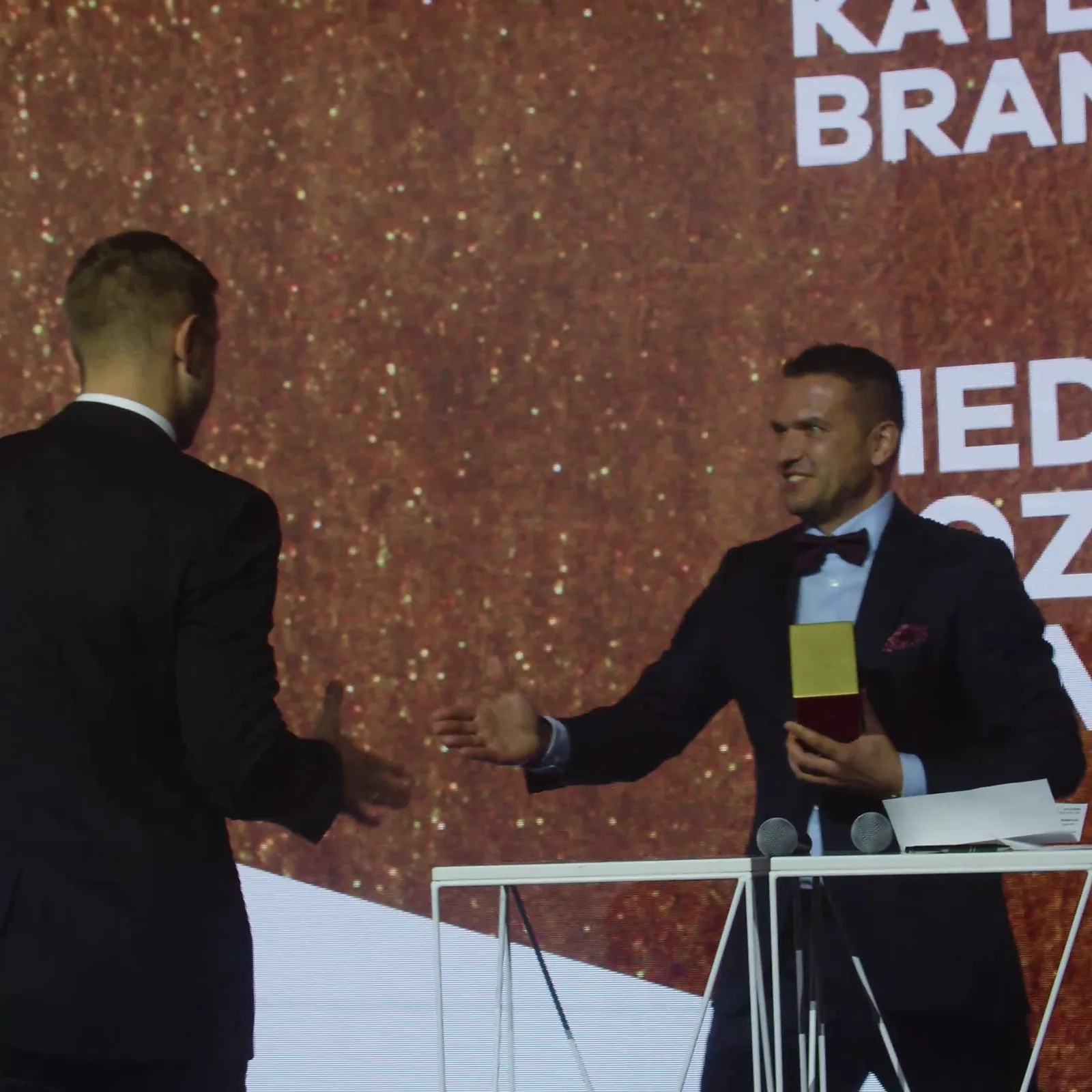 Prowadzący wyciąga dłoń do laureata, który wchodzi na scenę po odbiór statuetki — Gala DMR XII (2023)