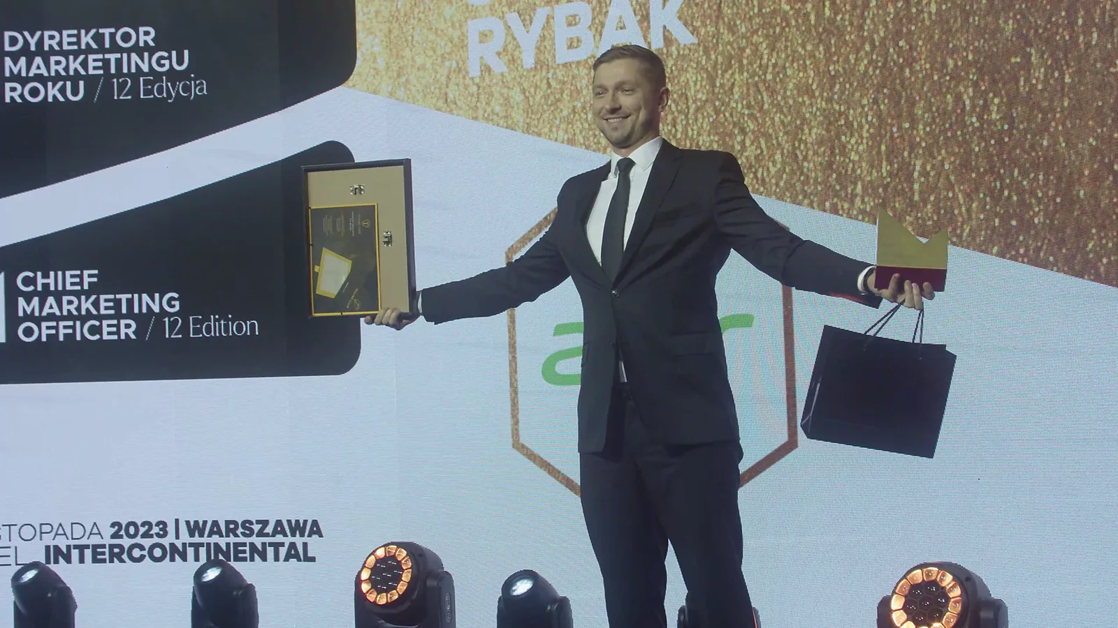 Laureat z rozłożonymi rękami prezentuje dyplom i torbę z upominkiem przed logo konkursu — Gala DMR XII (2023)
