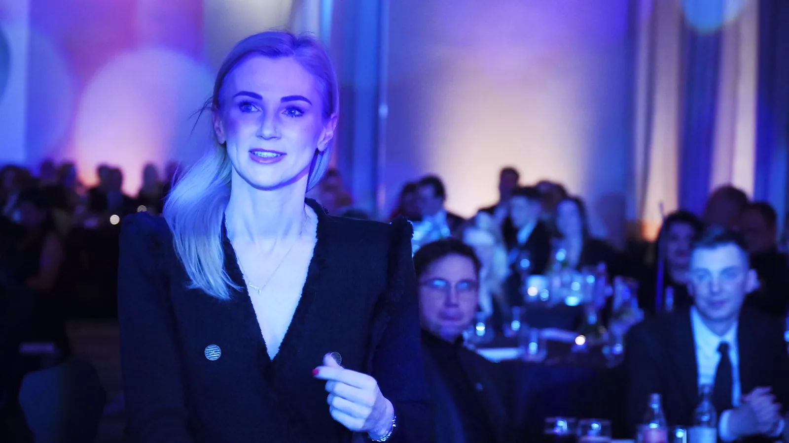 Uśmiechnięta blondynka w czarnej marynarce przechodzi przez salę pełną gości w niebieskim świetle — Gala DMR XII (2023)