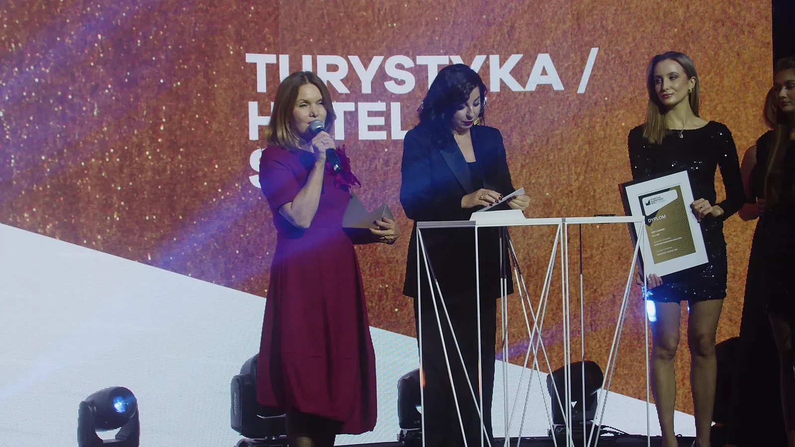 Kobieta w czerwonej sukience przemawia w kategorii turystyka i hotele, obok laureatka z dyplomem — Gala DMR XII (2023)