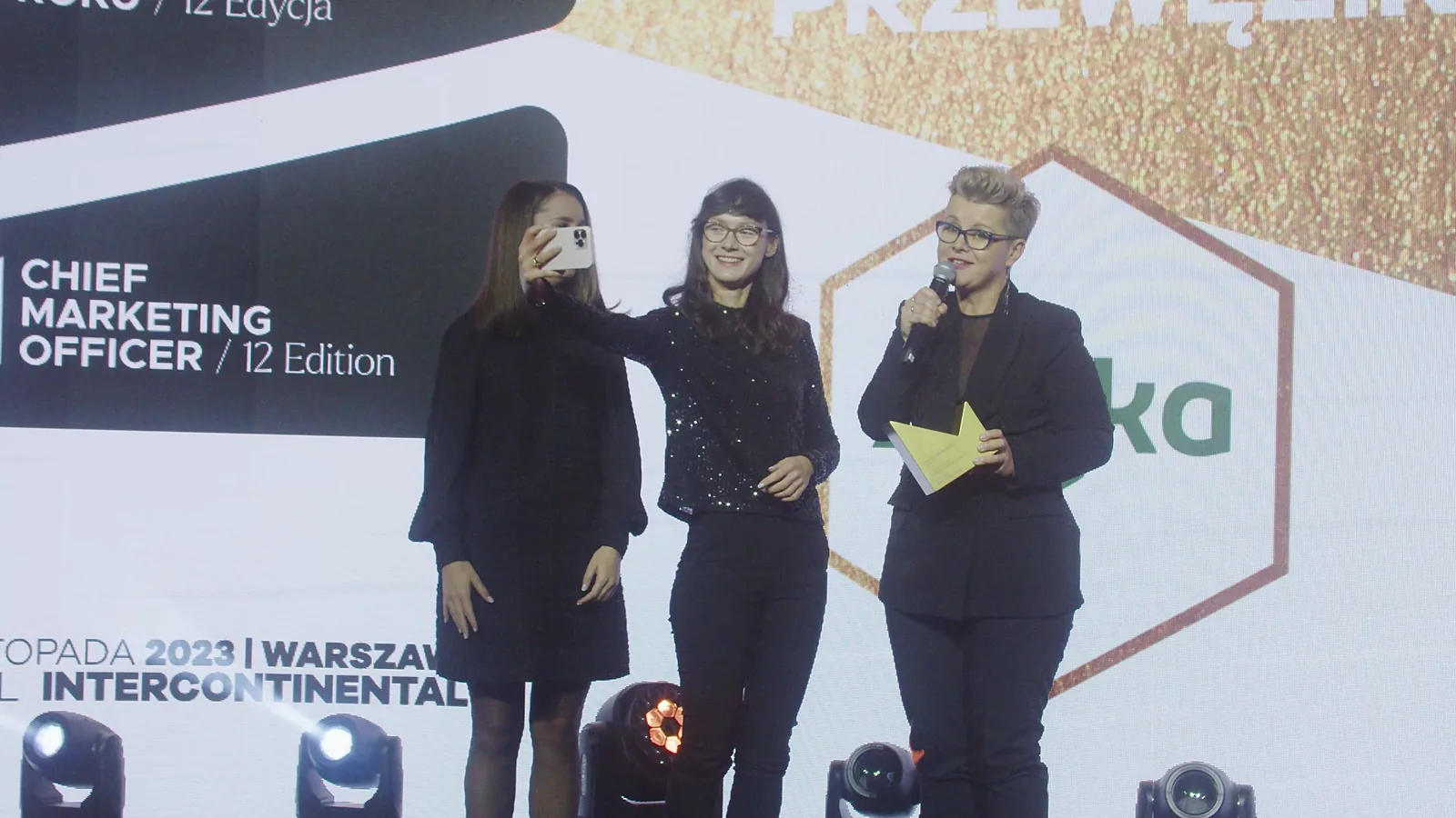 Trzy laureatki na scenie: jedna mówi do mikrofonu, druga robi zdjęcie telefonem — Gala DMR XII (2023)