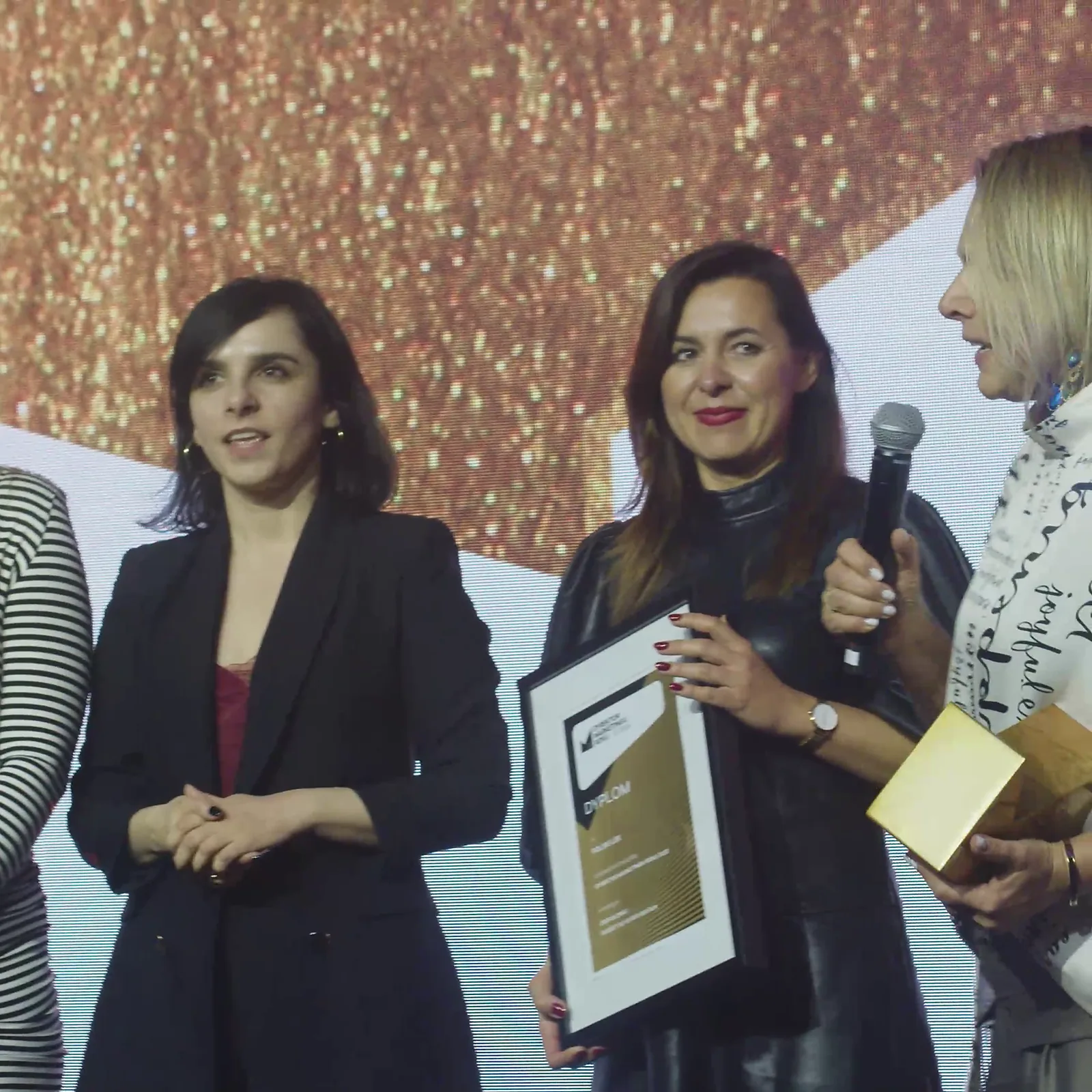 Three women on stage: a winner with a diploma and microphone, a colleague holding the statuette — DMR Gala XII (2023)