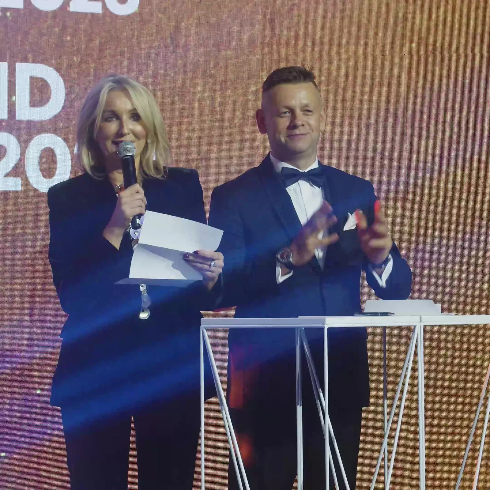 Duet prowadzących przy białej mównicy ogłasza Grand Prix, prowadzący klaszcze w dłonie — Gala DMR XII (2023)