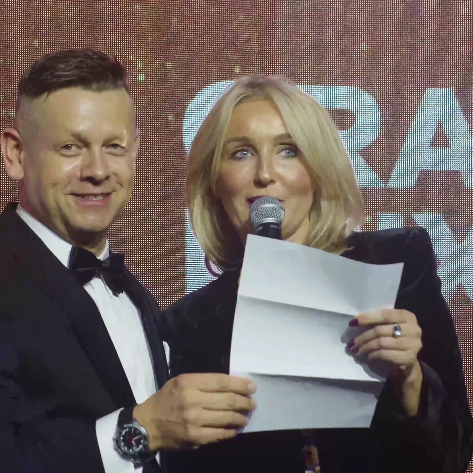 Zbliżenie: prowadząca czyta z kartki werdykt Grand Prix, a prowadzący szeroko się uśmiecha — Gala DMR XII (2023)