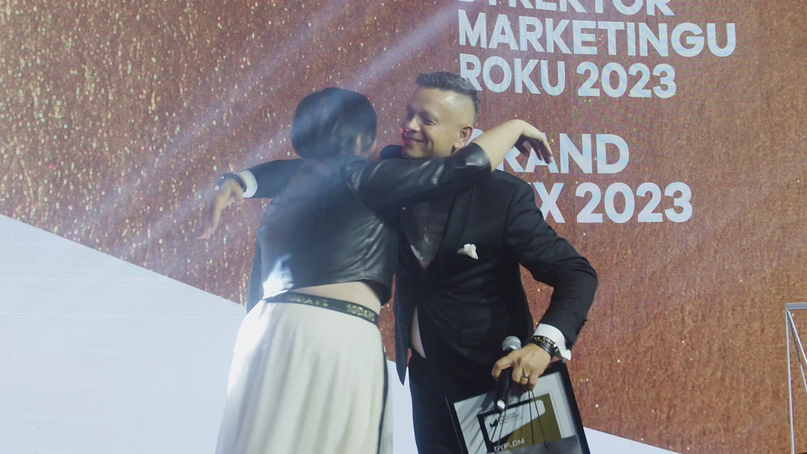 Laureatka w białej spódnicy obejmuje prowadzącego, który trzyma jej oprawiony dyplom — Gala DMR XII (2023)