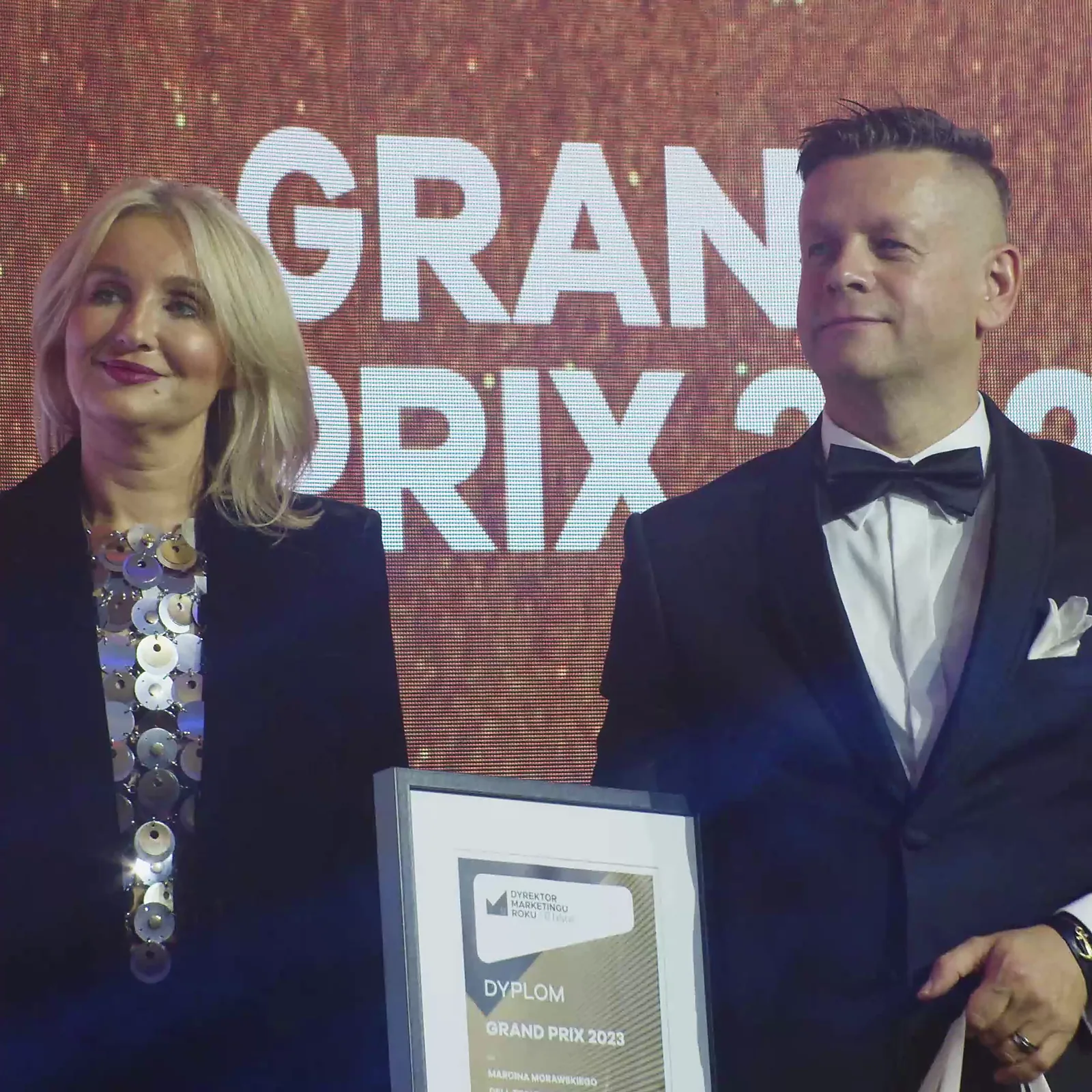 Kobieta w ciemnym żakiecie z dyplomem Grand Prix i uśmiechnięty prowadzący w smokingu — Gala DMR XII (2023)