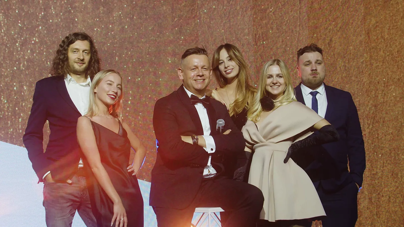 Pięcioro gości w wieczorowych strojach pozuje z siedzącym prowadzącym na tle złotej ściany — Gala DMR XII (2023)